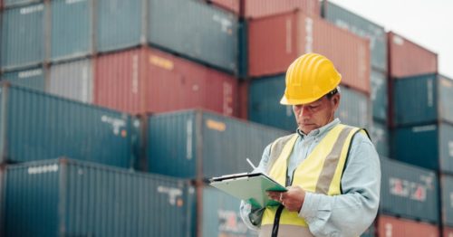 Operario revisando documentación aduanera junto a contenedores en una terminal portuaria